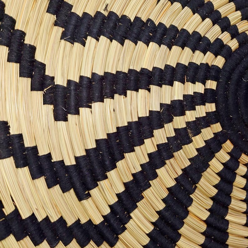 May include: Close-up of a woven basket with a repeating geometric pattern. The basket features alternating bands of natural beige and black fibres, creating a striking visual contrast. The design is circular, with a central spiral.