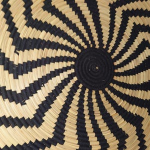 May include: Close-up of a woven basket with a spiral design. The basket features alternating bands of natural beige and black, creating a striking geometric pattern. The centre is a solid black circle.