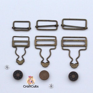 May include: Assortment of antique gold metal garment clasps and snaps. The clasps are in three sizes and have a rectangular shape with a small loop on each side. The snaps are round and have a star design.
