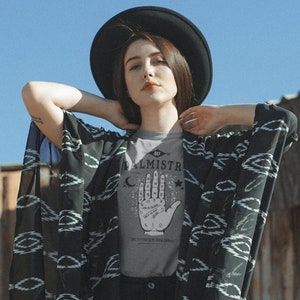 May include: A woman wearing a grey t-shirt with a black and white illustration of a hand with lines and symbols. The text "The Art of Palmistry" and "The future is in your hands" are printed on the shirt. She is wearing a black hat and a black and white patterned kimono.