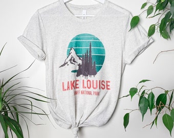 May include: Heather gray t-shirt with a graphic of a mountain, trees, and a teal and green circle. The text "Lake Louise Banff National Park" is printed below the graphic. The shirt is hanging on a white hanger.