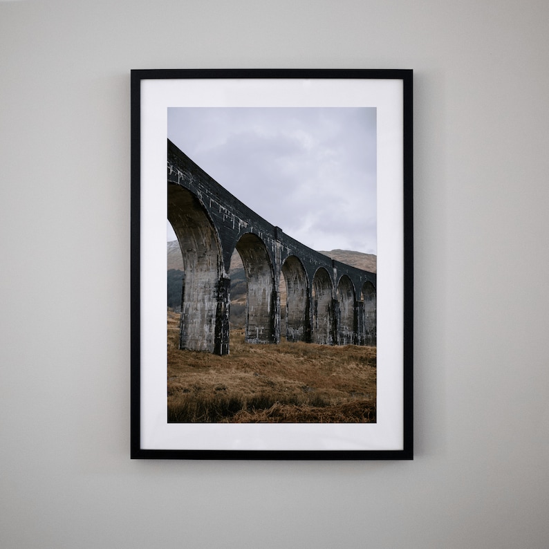 May include: A black and white photograph of a stone arch bridge in a rural landscape. The bridge is made of large, rough-hewn stones and has a series of arches that span a valley. The bridge is surrounded by green grass and trees.