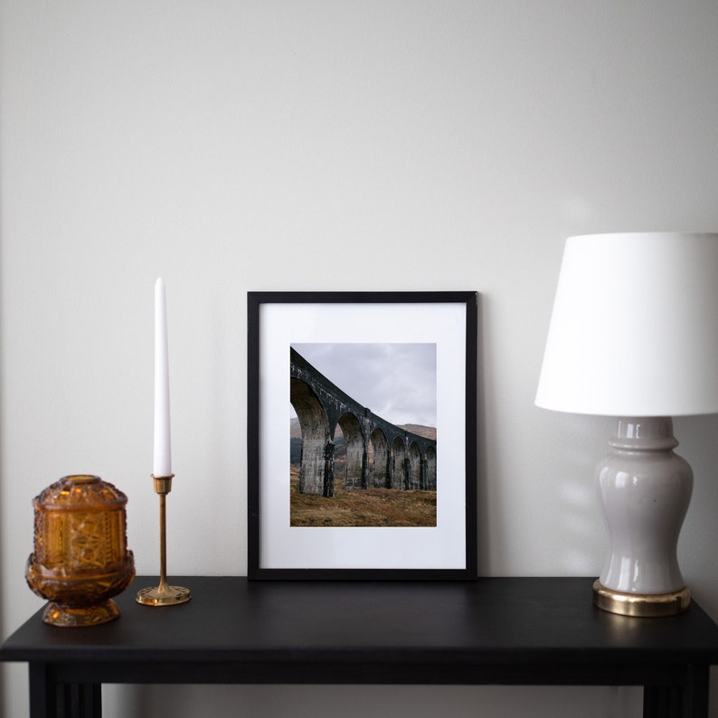 May include: A black framed photograph of a stone arch bridge with a cloudy sky in the background. The bridge is made of stone and has a series of arches. The photograph is on a black table with a candle holder and a lamp.
