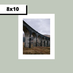 May include: A black stone arch bridge with multiple arches spanning a valley. The bridge is made of concrete and has a rough texture. The bridge is surrounded by green grass and trees.
