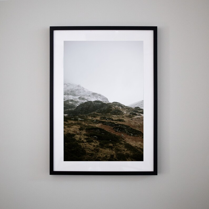 May include: A framed photograph of a mountain range with snow-capped peaks and green slopes. The photo is in a black frame and is hanging on a white wall.