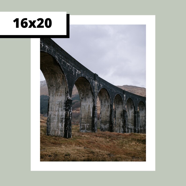 May include: A black stone arch bridge with multiple arches spanning a valley. The bridge is made of large, rough-hewn stones and has a rustic, weathered appearance. The bridge is set against a backdrop of rolling hills and a cloudy sky.