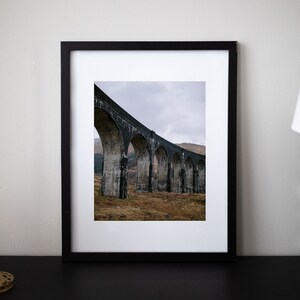 May include: A black framed photograph of a stone arch bridge in a rural landscape. The bridge is made of large, rough-hewn stones and has multiple arches. The bridge is set against a backdrop of rolling hills and a cloudy sky.