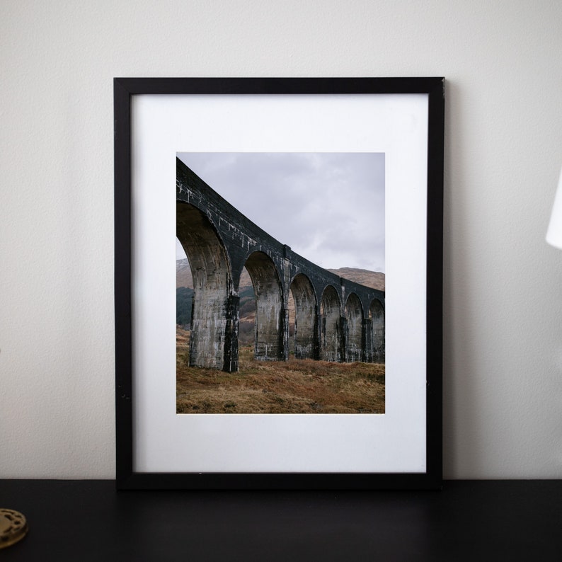 May include: A black framed photograph of a stone arch bridge in a rural landscape. The bridge is made of large, rough-hewn stones and has multiple arches. The bridge is set against a backdrop of rolling hills and a cloudy sky.