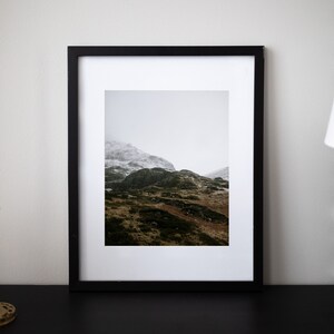 May include: A black framed photograph of a mountain range with snow-capped peaks and green slopes. The photo is taken on a cloudy day.