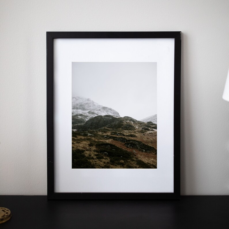 May include: A black framed photograph of a mountain range with snow-capped peaks and green slopes. The photo is taken on a cloudy day.