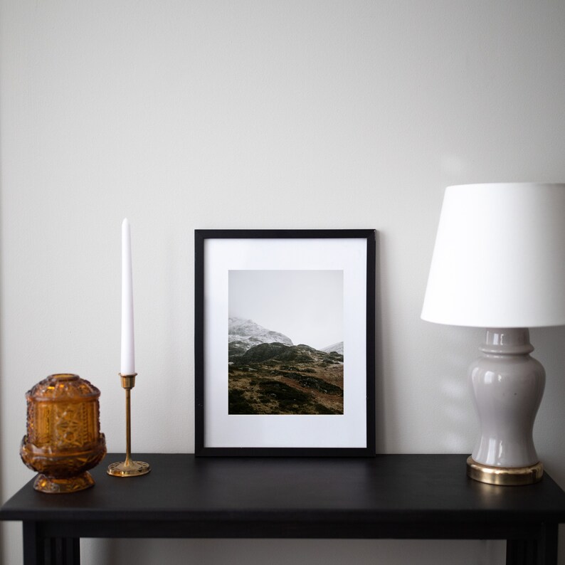 May include: A black framed photo of a mountain landscape with snow on the peaks. The photo is on a black table with a gold candle holder and a white candle, and a white lamp with a gray base.