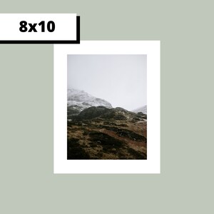 May include: A photograph of a mountain range with snow-capped peaks and green slopes. The sky is overcast and the mountains are shrouded in mist.