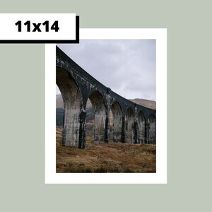 May include: A black stone arch bridge with multiple arches spanning a valley. The bridge is made of large, rough-hewn stones and is set against a backdrop of green hills.