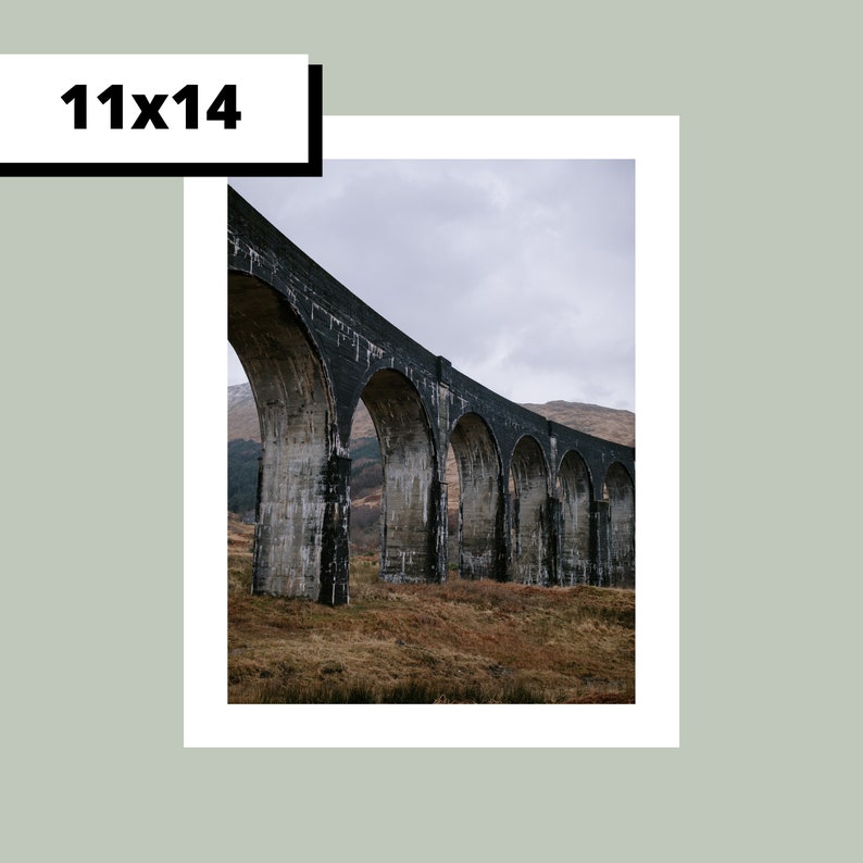 May include: A black stone arch bridge with multiple arches spanning a valley. The bridge is made of large, rough-hewn stones and is set against a backdrop of green hills.