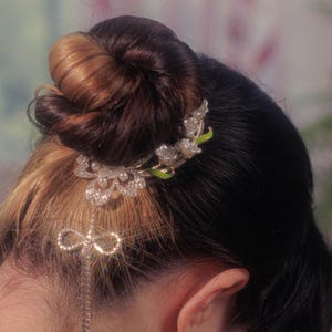 May include: A close-up of a hairstyle featuring a bun with a decorative hair clip. The clip is silver-toned with a floral design and dangling embellishments. The hair is a mix of dark brown and light brown.