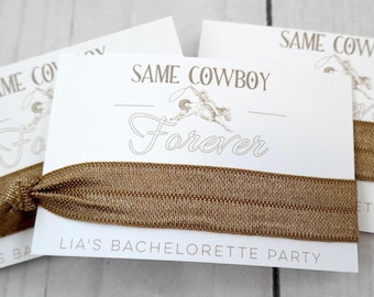 May include: Two white cards with the text "Same Cowboy Forever" and "Lia's Bachelorette Party" printed on them. A brown elastic hair tie is wrapped around the cards.