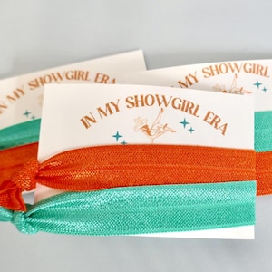 May include: Three hair tie cards with the text "IN MY SHOWGIRL ERA" in brown. Each card has a different colored hair tie: orange and teal. The cards have a vintage showgirl illustration and star accents.