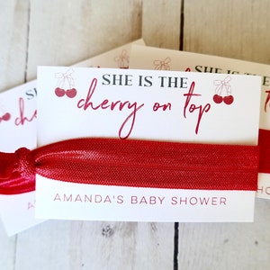May include: White card with red text that reads "She is the cherry on top" with a red cherry graphic. The card is attached to a red elastic hair tie. The text "Amanda's Baby Shower" is printed on the bottom of the card.