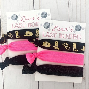 May include: Two sets of hair ties with cards that read "Lana's LAST RODEO". Each set includes a black tie with gold cowboy boot and hat designs, a bright pink tie, and a black glitter tie.