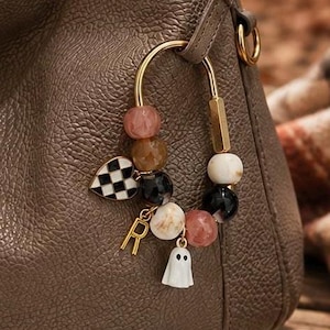 May include: A taupe textured leather handbag featuring a gold-toned U-shaped carabiner clip. The clip is adorned with a collection of beads in shades of pink, brown, black, and white, a black and white checkered heart charm, a gold letter R, and a white ghost.