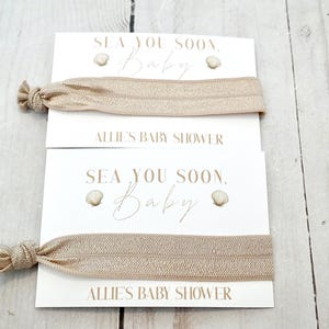 May include: Two beige elastic hair ties are displayed on white cards. The cards read "SEA YOU SOON, Baby" with seashell graphics and "ALLIE'S BABY SHOWER" in a delicate font. The background is a light wood surface.