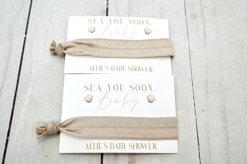 May include: Two beige elastic hair ties are displayed on white cards. The cards read "SEA YOU SOON, Baby" with seashell graphics and "ALLIE'S BABY SHOWER" in a delicate font. The background is a light wood surface.