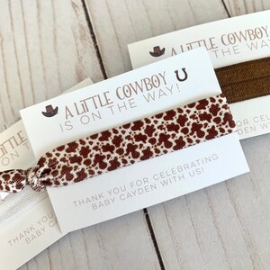 May include: A white card with brown text that reads "A Little Cowboy is on the way!" with a brown cowboy hat icon. A brown and white patterned hair tie is attached to the card. The card also has the text "Thank you for celebrating Baby Cayden with us!"