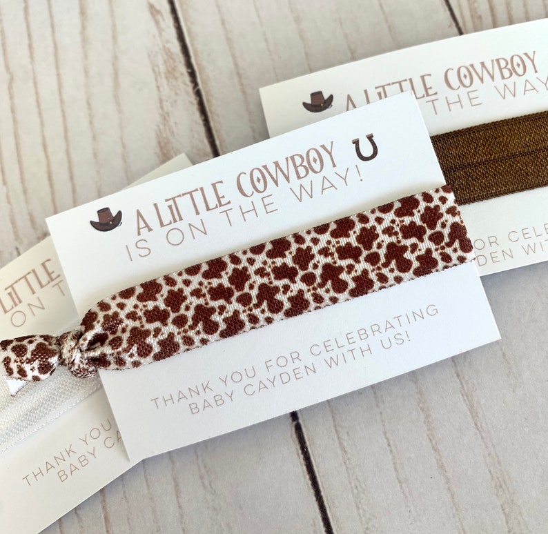 May include: A white card with brown text that reads "A Little Cowboy is on the way!" with a brown cowboy hat icon. A brown and white patterned hair tie is attached to the card. The card also has the text "Thank you for celebrating Baby Cayden with us!"