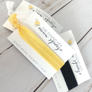 May include: Three white cards with black text and a lemon graphic. The text reads "She Found Her Main Squeeze". The cards are attached to yellow and black elastic hair ties.