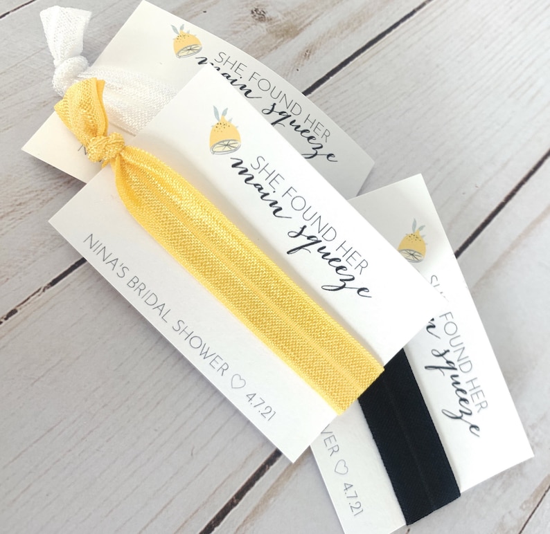 May include: Three white cards with black text and a lemon graphic. The text reads "She Found Her Main Squeeze". The cards are attached to yellow and black elastic hair ties.