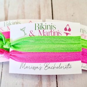 May include: Two green and pink hair ties with a white card that says "Bikinis & Martinis" and "Marissa's Bachelorette" in black cursive writing. The card also has a martini glass and a bikini graphic.