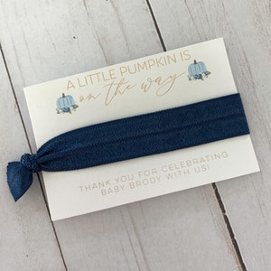 May include: A white card with blue and orange pumpkins and the text "A little pumpkin is on the way". A navy blue hair tie is attached to the card. The card also says "Thank you for celebrating baby Brody with us!"