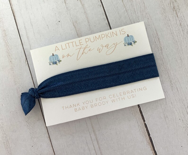 May include: A white card with blue and orange pumpkins and the text "A little pumpkin is on the way". A navy blue hair tie is attached to the card. The card also says "Thank you for celebrating baby Brody with us!"
