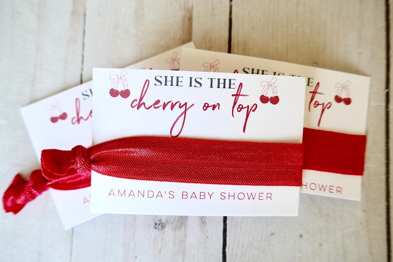 May include: White card with red text that reads "She is the cherry on top" with a red cherry graphic. The card is attached to a red elastic hair tie. The text "Amanda's Baby Shower" is printed on the bottom of the card.