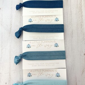 May include: Four blue and light blue hair ties with a white card attached to each. The card has a blue and white pumpkin illustration and the text "A little pumpkin is on the way". The card also includes the text "Thank you for celebrating baby Brody with us!"