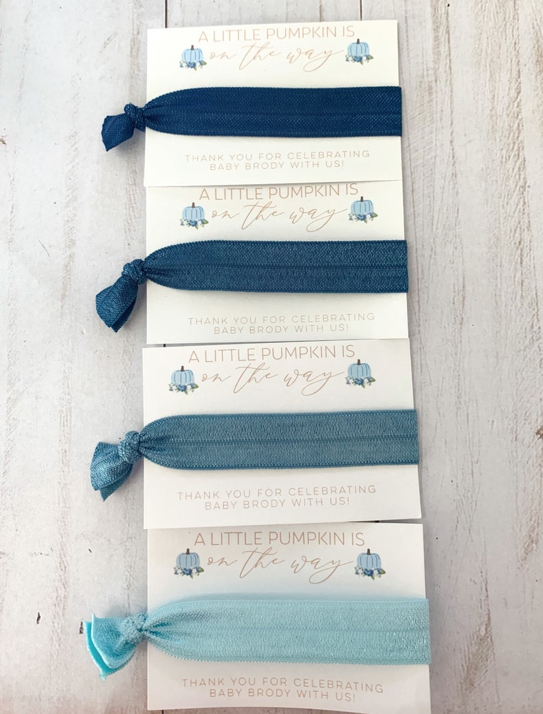 May include: Four blue and light blue hair ties with a white card attached to each. The card has a blue and white pumpkin illustration and the text "A little pumpkin is on the way". The card also includes the text "Thank you for celebrating baby Brody with us!"