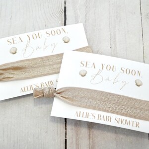 May include: Two baby shower favors featuring a white card with the text "SEA YOU SOON. Baby" and a seashell graphic. A beige elastic hair tie is attached. The card also reads "ALLIE'S BABY SHOWER".