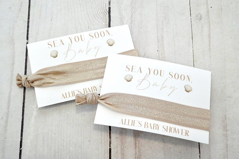 May include: Two baby shower favors featuring a white card with the text "SEA YOU SOON. Baby" and a seashell graphic. A beige elastic hair tie is attached. The card also reads "ALLIE'S BABY SHOWER".