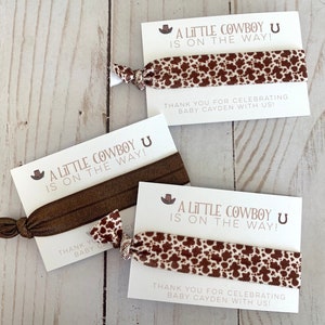 May include: Three brown and white patterned hair ties with a white card attached to each. The card says "A Little Cowboy U Is On The Way!" and "Thank You For Celebrating Baby Cayden With Us!"