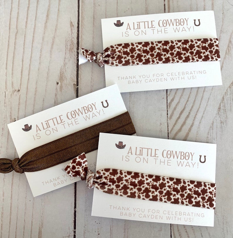 May include: Three brown and white patterned hair ties with a white card attached to each. The card says "A Little Cowboy U Is On The Way!" and "Thank You For Celebrating Baby Cayden With Us!"
