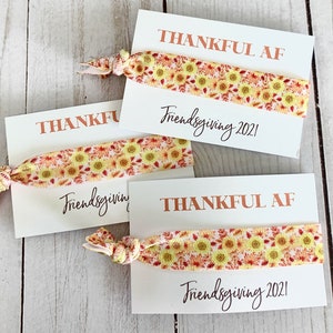 May include: Three white cards with the text "THANKFUL AF" and "Friendsgiving 2021" printed in brown and black. Each card has a sunflower patterned hair tie. The hair ties are yellow, orange, and red.