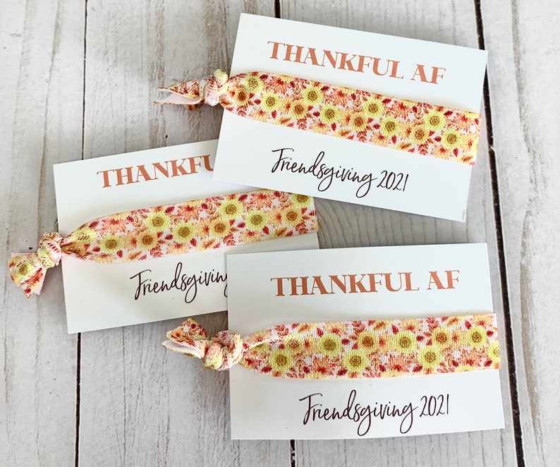 May include: Three white cards with the text "THANKFUL AF" and "Friendsgiving 2021" printed in brown and black. Each card has a sunflower patterned hair tie. The hair ties are yellow, orange, and red.