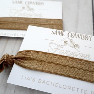 K&ouml;nnte beinhalten: Eine wei&szlig;e Karte mit dem Text "Same Cowboy Forever" und "Lia's Bachelorette Party" darauf gedruckt. Ein braunes Gummiband ist um die Karte gewickelt.