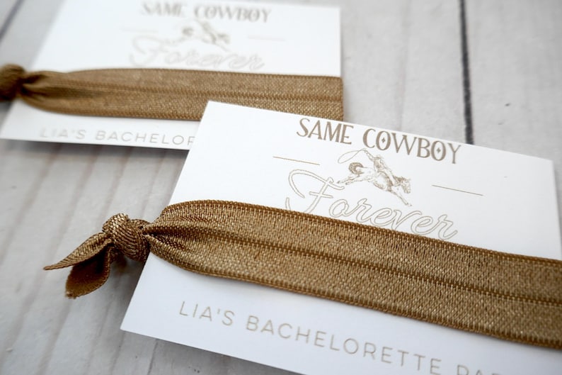 K&ouml;nnte beinhalten: Eine wei&szlig;e Karte mit dem Text "Same Cowboy Forever" und "Lia's Bachelorette Party" darauf gedruckt. Ein braunes Gummiband ist um die Karte gewickelt.