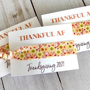 May include: Three white cards with the words "THANKFUL AF" printed in copper-colored ink. Each card holds a hair tie with a yellow sunflower pattern. The words "Friendsgiving 2021" are written in black script.