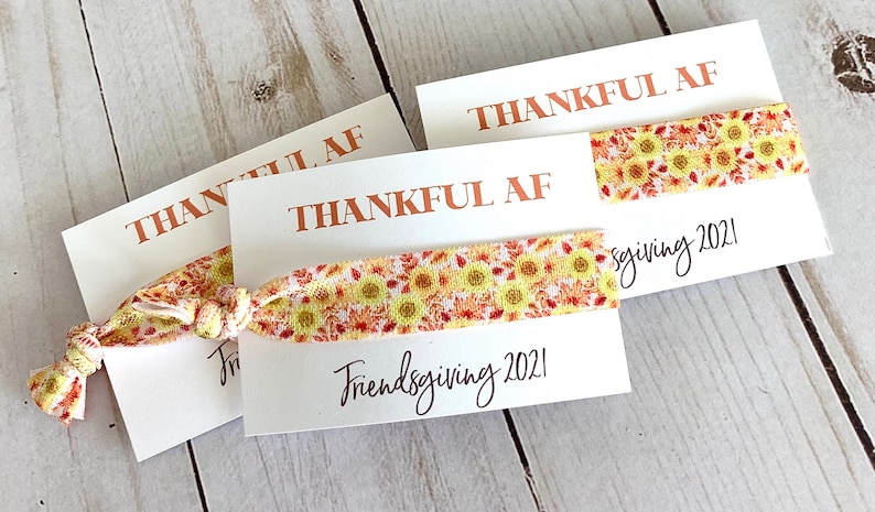 May include: Three white cards with the words "THANKFUL AF" printed in copper-colored ink. Each card holds a hair tie with a yellow sunflower pattern. The words "Friendsgiving 2021" are written in black script.
