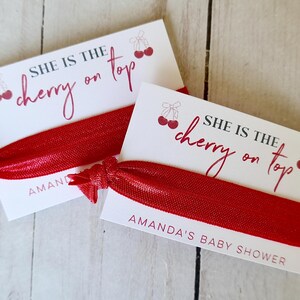 May include: Two white cards with red cherries and the text "She is the cherry on top" with red hair ties. The cards have the text "Amanda's Baby Shower" printed on them.
