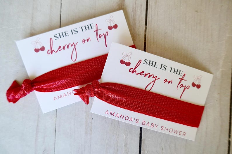 May include: Two white cards with red cherries and the text "She is the cherry on top" with red hair ties. The cards have the text "Amanda's Baby Shower" printed on them.