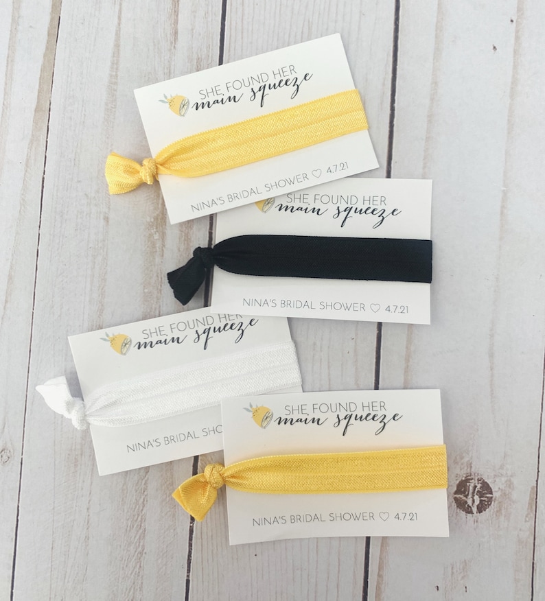May include: Four cards with lemon illustrations and the text "She found her main squeeze" with a yellow, black, white, and yellow hair tie on each card. The cards also include the text "Nina's Bridal Shower 4.7.21".