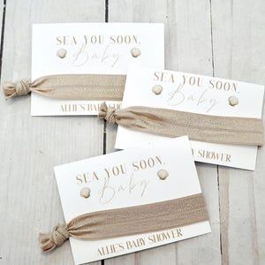 May include: Three white cards with the text "SEA YOU SOON, Baby" and a seashell graphic. Each card has a tan elastic hair tie attached. The text "ALLIE'S BABY SHOWER" is printed on the bottom of one card.